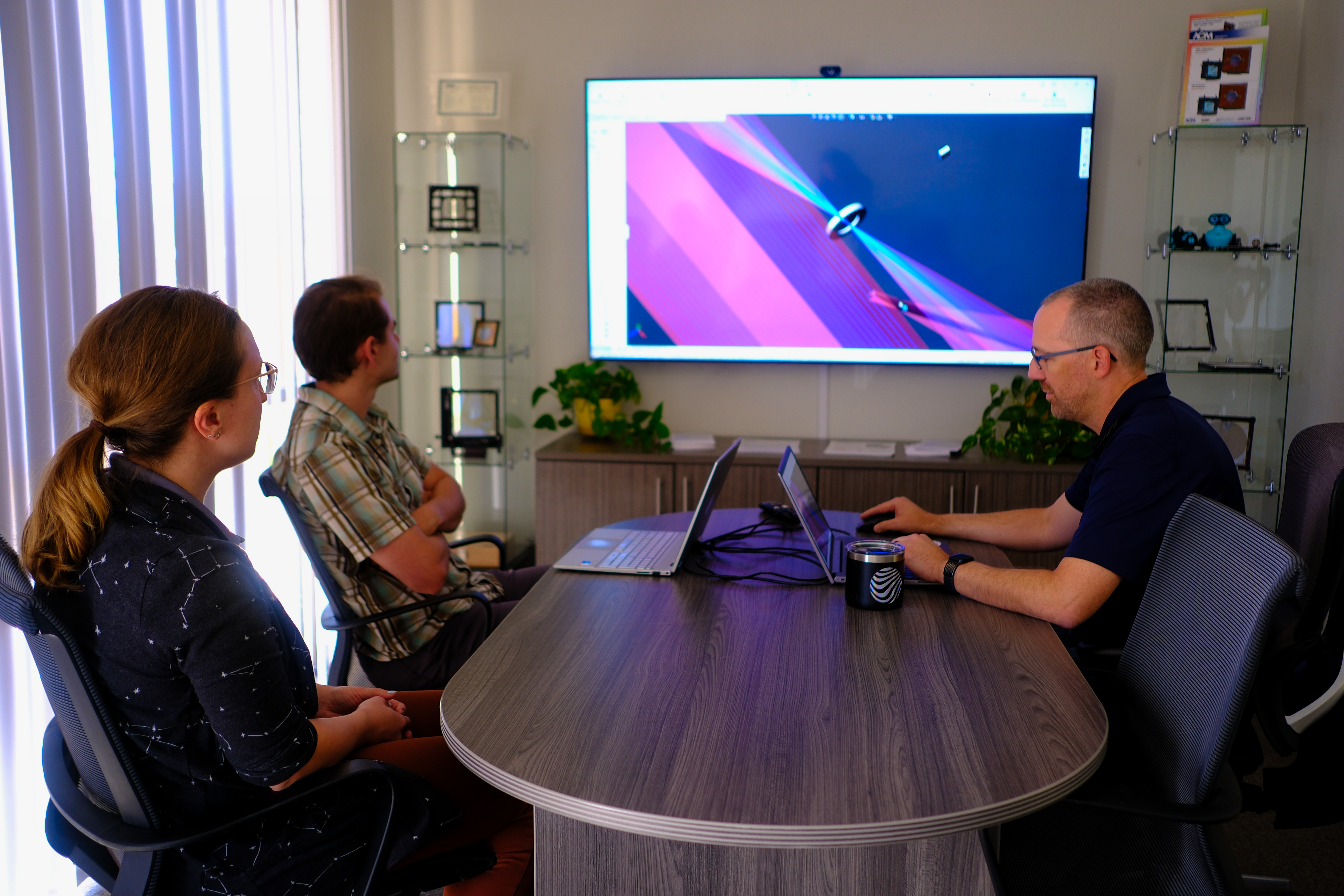 Three engineers sitting at a conference table analyze a ray diagram of an optical system on a large TV screen